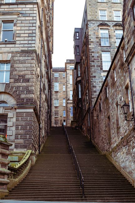 A narrow stone passageway inside a historic building in Edinburgh Old Town, with a view towards the city's skyline including a prominent Gothic-style church spire. The archway is dark, with stone wall